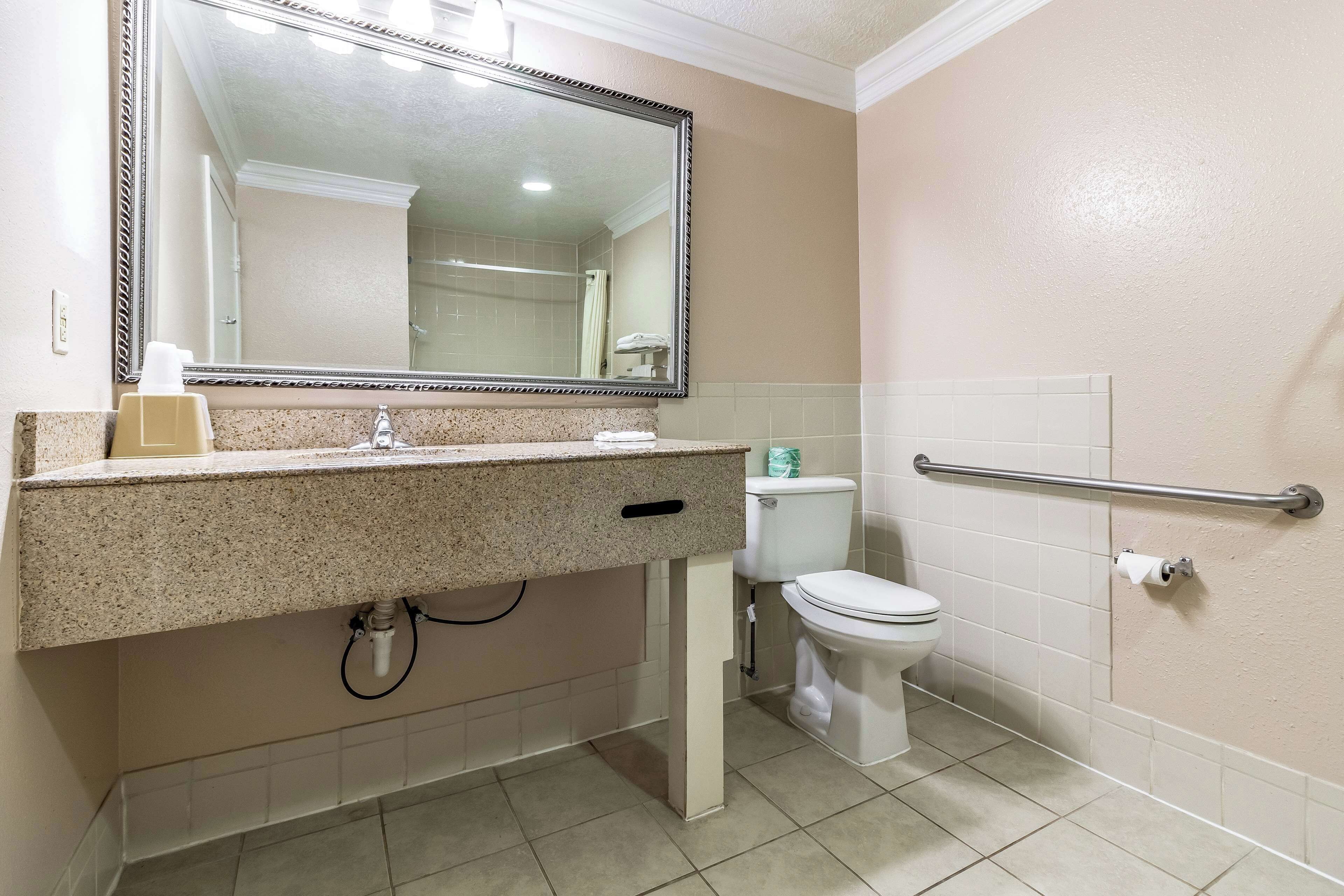 A bathroom with a granite countertop, a white toilet, a handrail, and a large mirror.