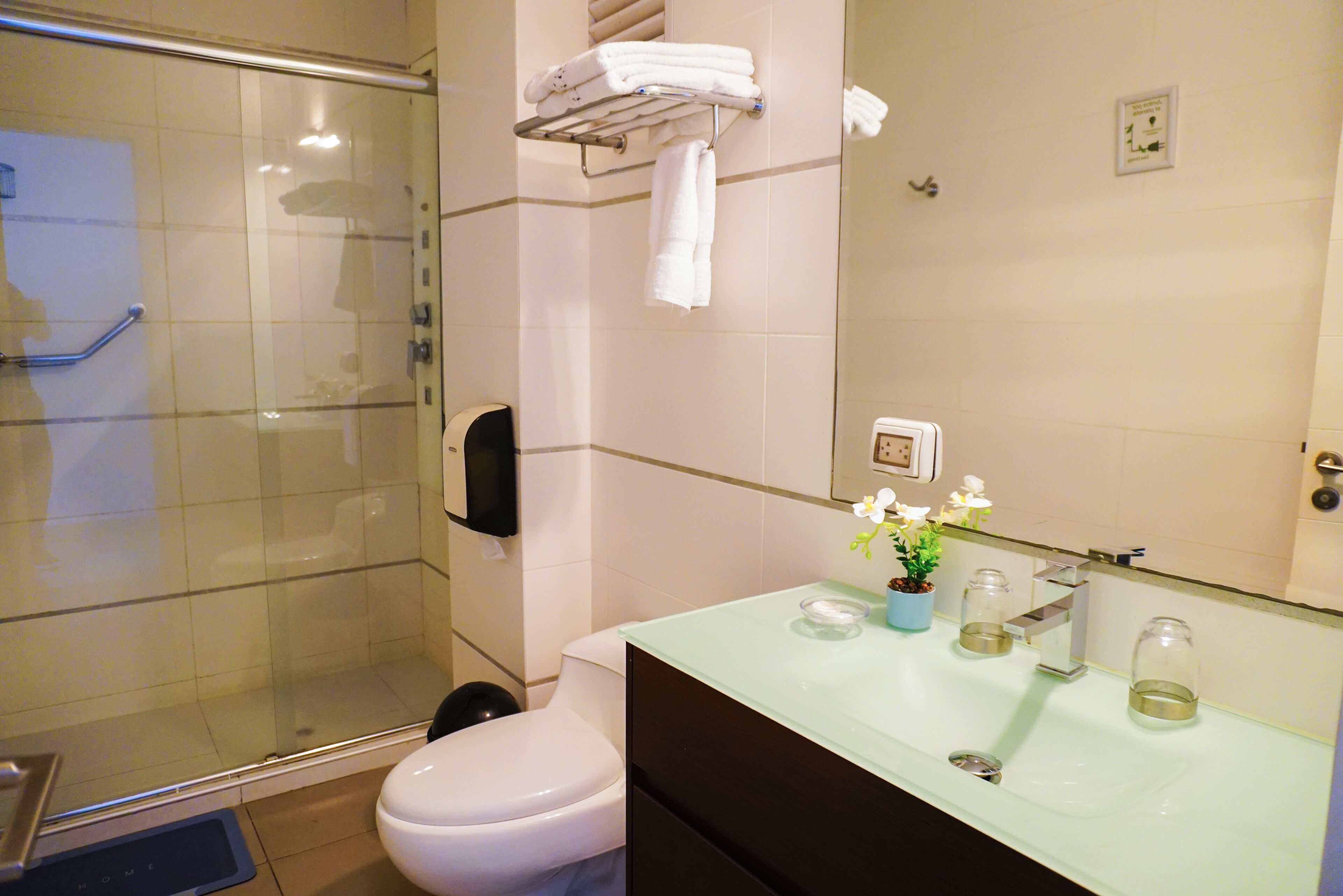 Un baño con cabina de ducha de vidrio, inodoro, lavamanos con encimera de vidrio y un perchero con toallas dobladas.