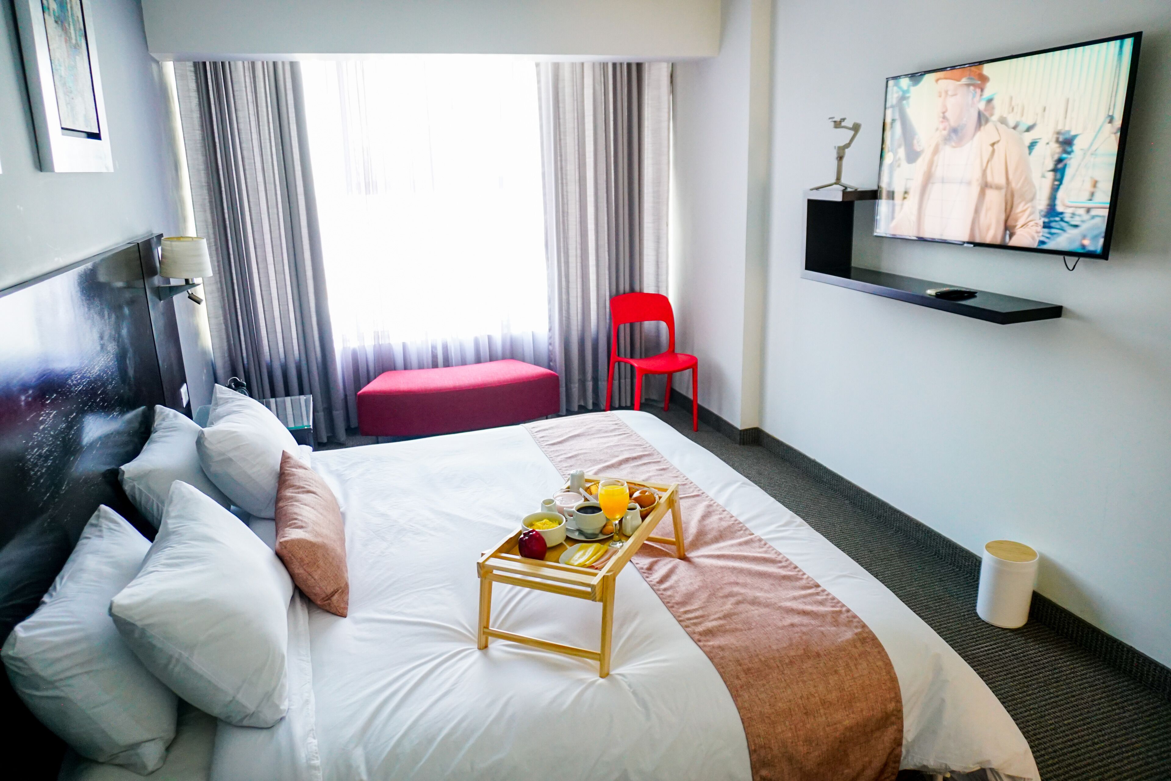 A hotel room with a bed, a TV, a breakfast tray, and a red chair.