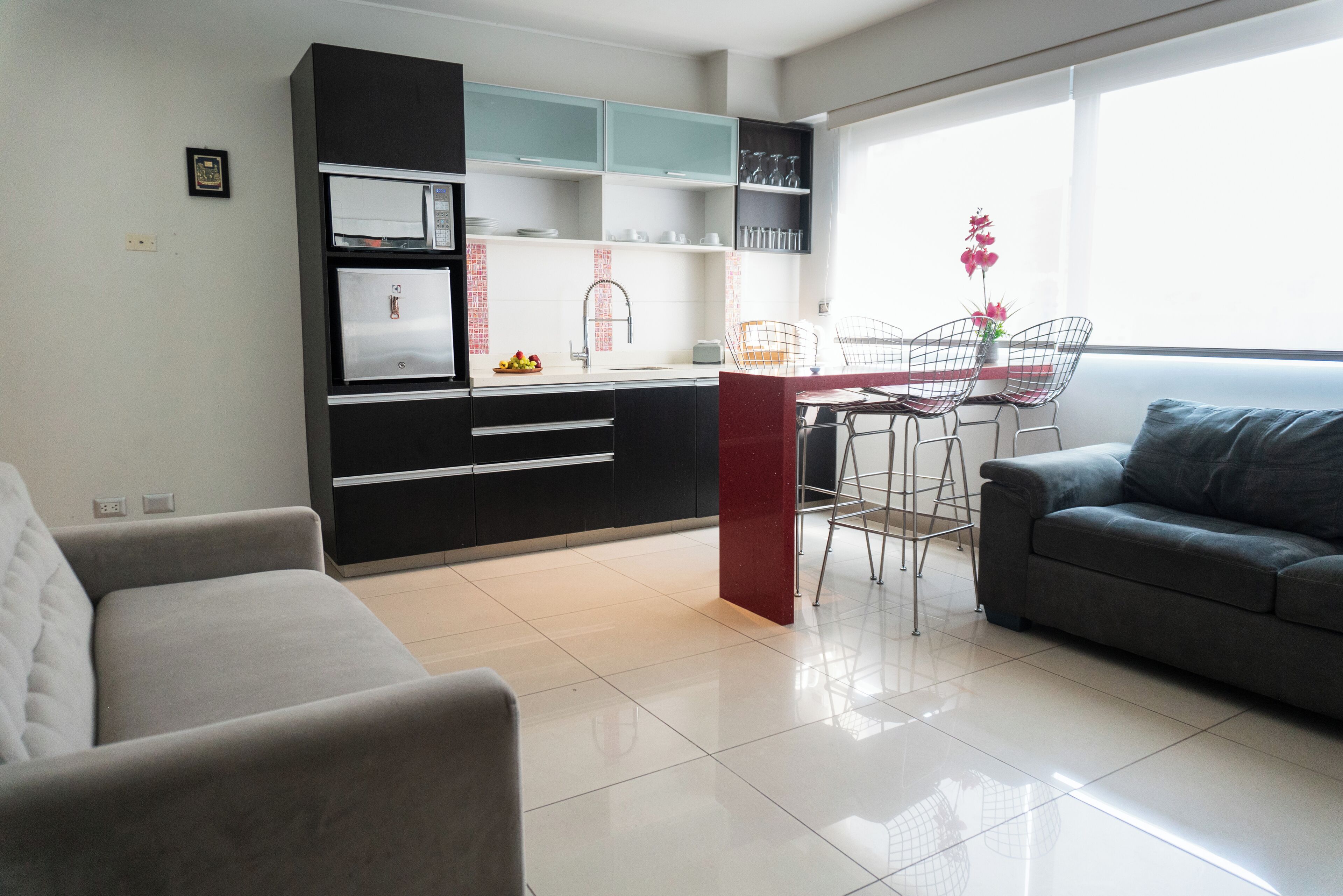 Una cocina moderna con armarios negros, encimera blanca e isla roja. Hay un comedor con mesa y sillas, y un sofá gris en la sala de estar.