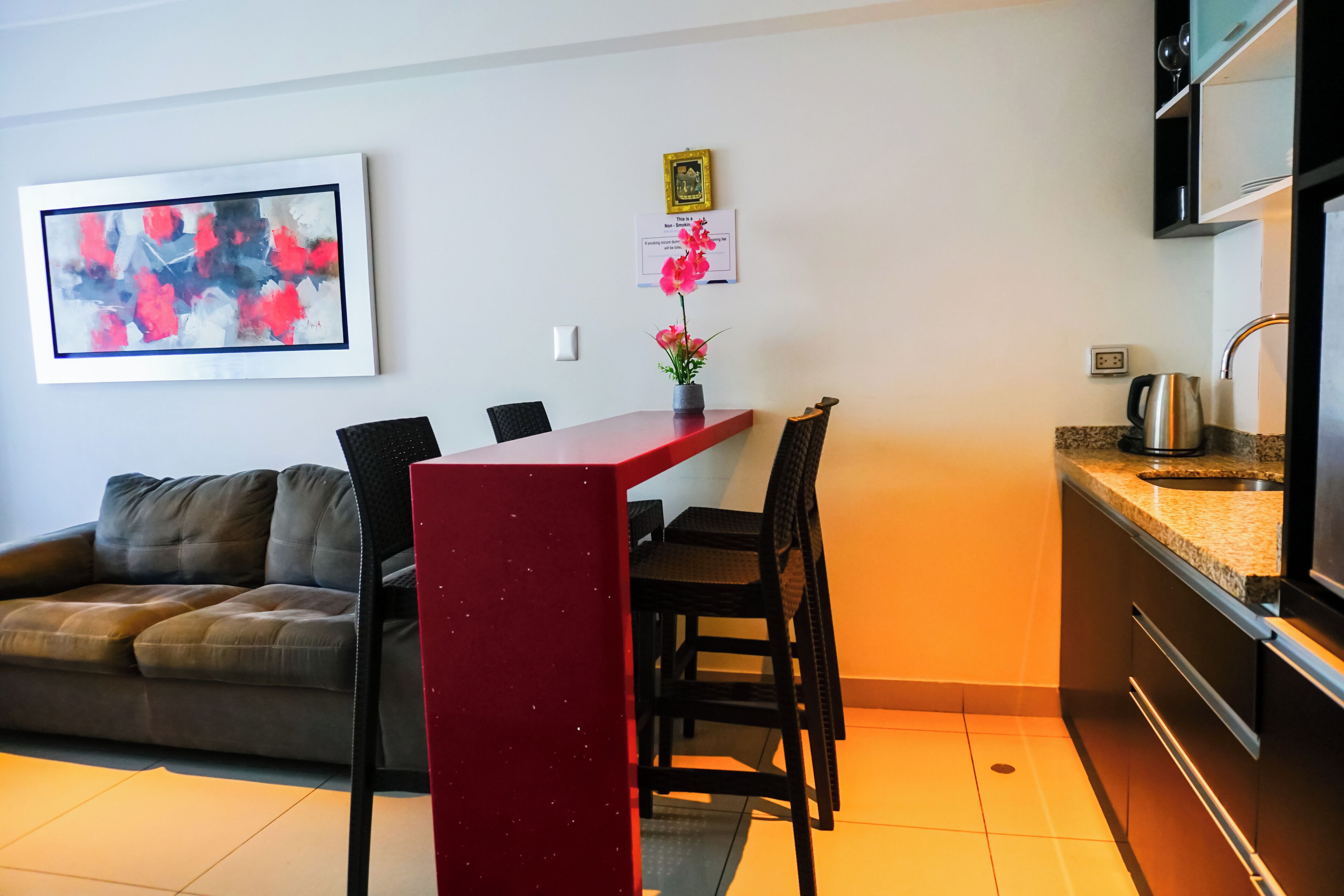 A compact living space with a kitchenette, a dining area with a red table and chairs, a sofa, and a framed abstract painting.