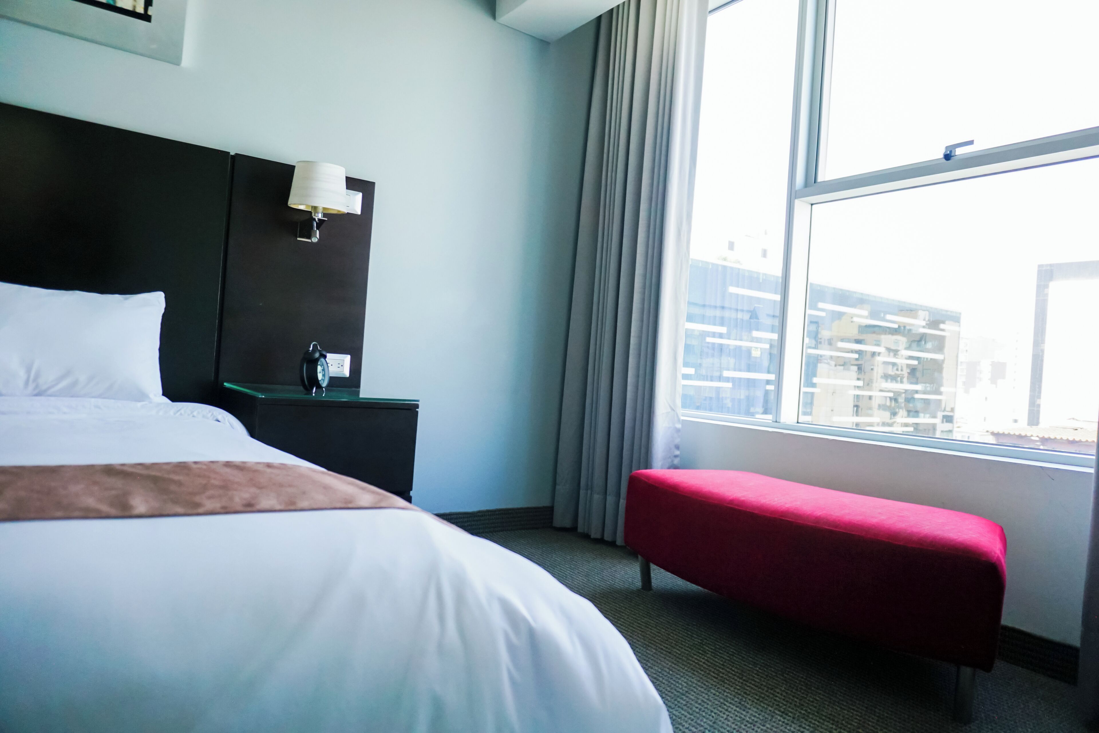 Une chambre d’hôtel avec un grand lit, une table de chevet, un banc rouge et une fenêtre donnant sur la ville.