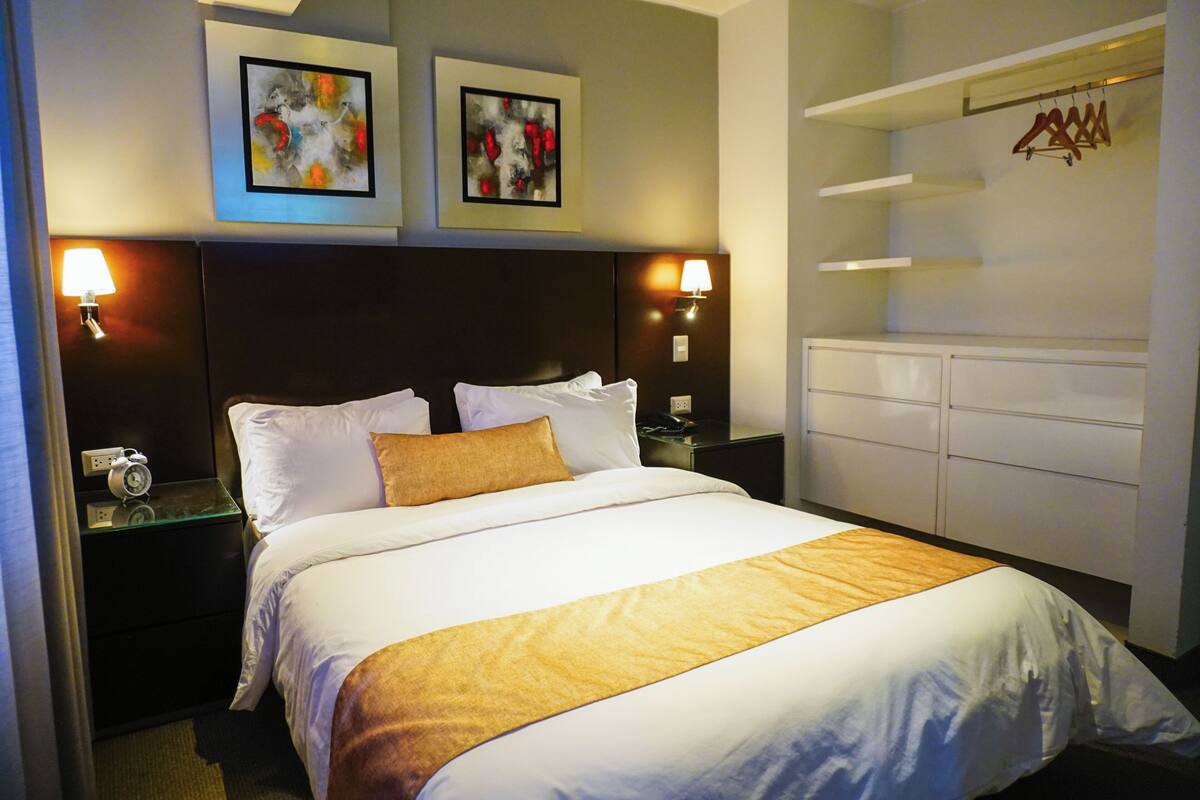 Quarto de hotel com uma cama grande, duas luminárias fixadas na parede, uma mesa de cabeceira com um relógio e um guarda-roupa com espaço para pendurar roupas.
