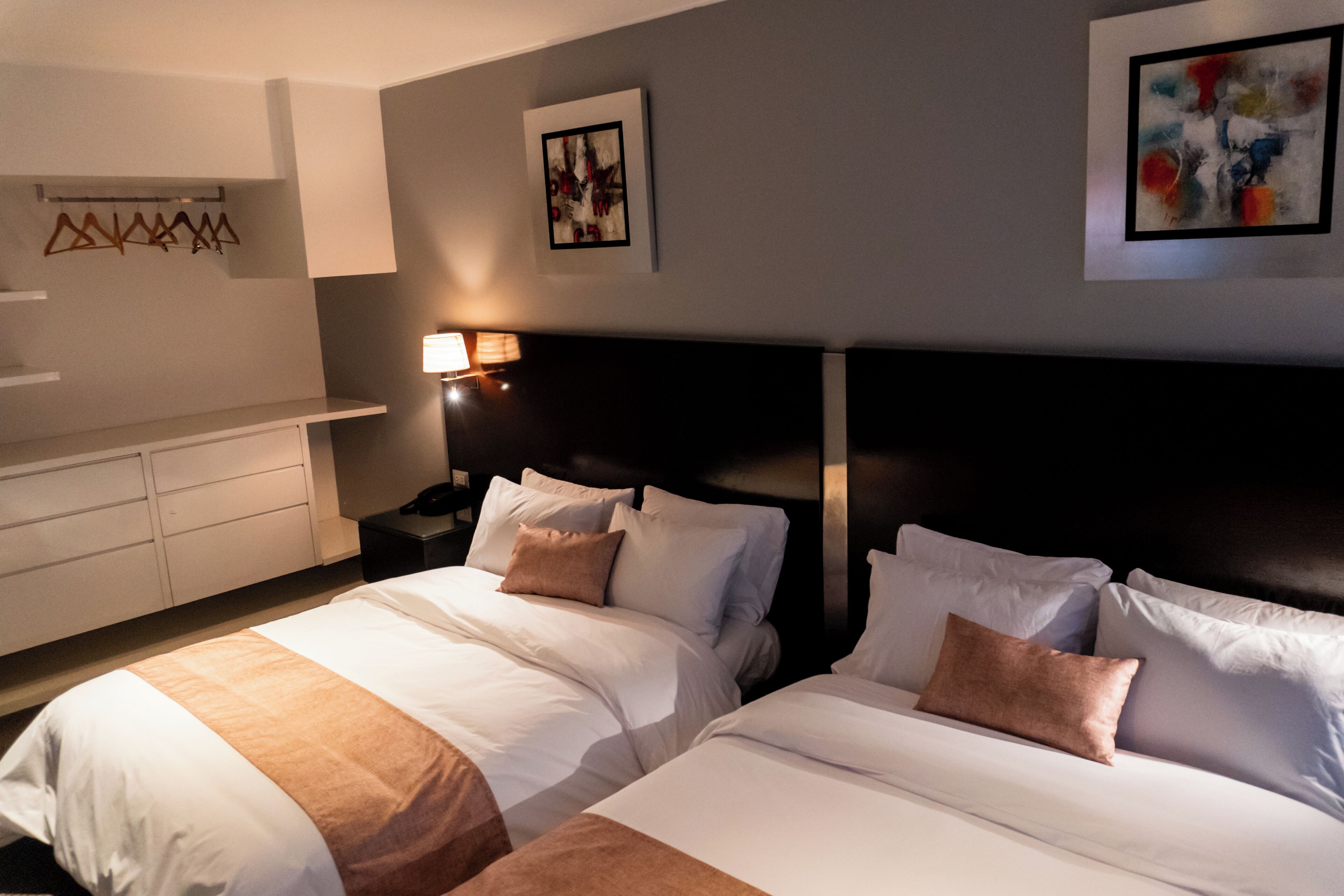A hotel room with two beds, a bedside table with a lamp, a wall-mounted coat rack, and artwork on the walls.