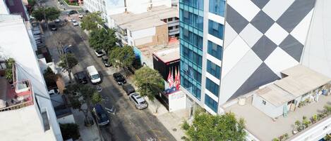 Un hotel de varios pisos con fachada moderna, un estacionamiento con coches y una vista de la calle con edificios y árboles.