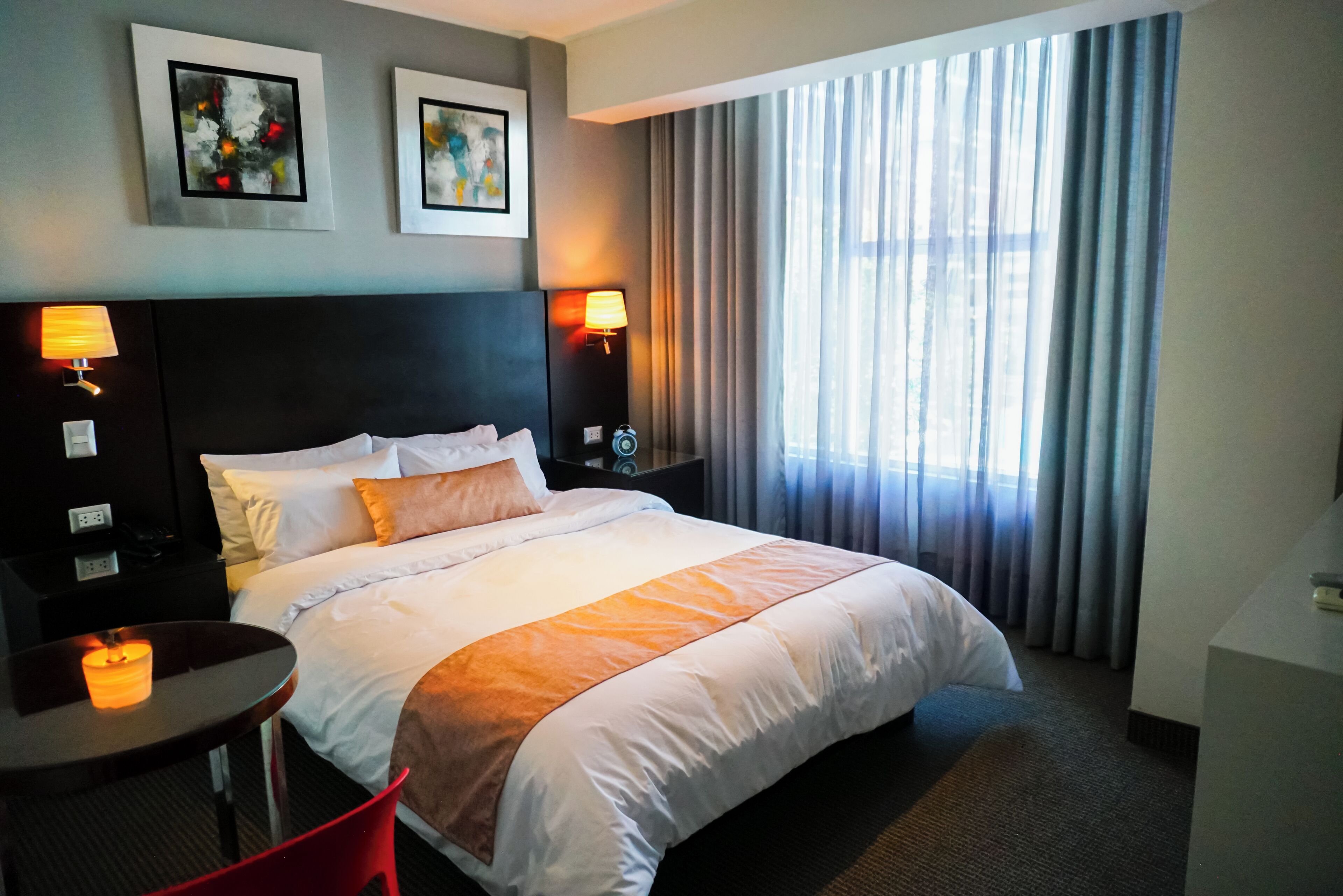 A hotel room with a large bed, two bedside tables with lamps, a small table with a candle, and a window with curtains.