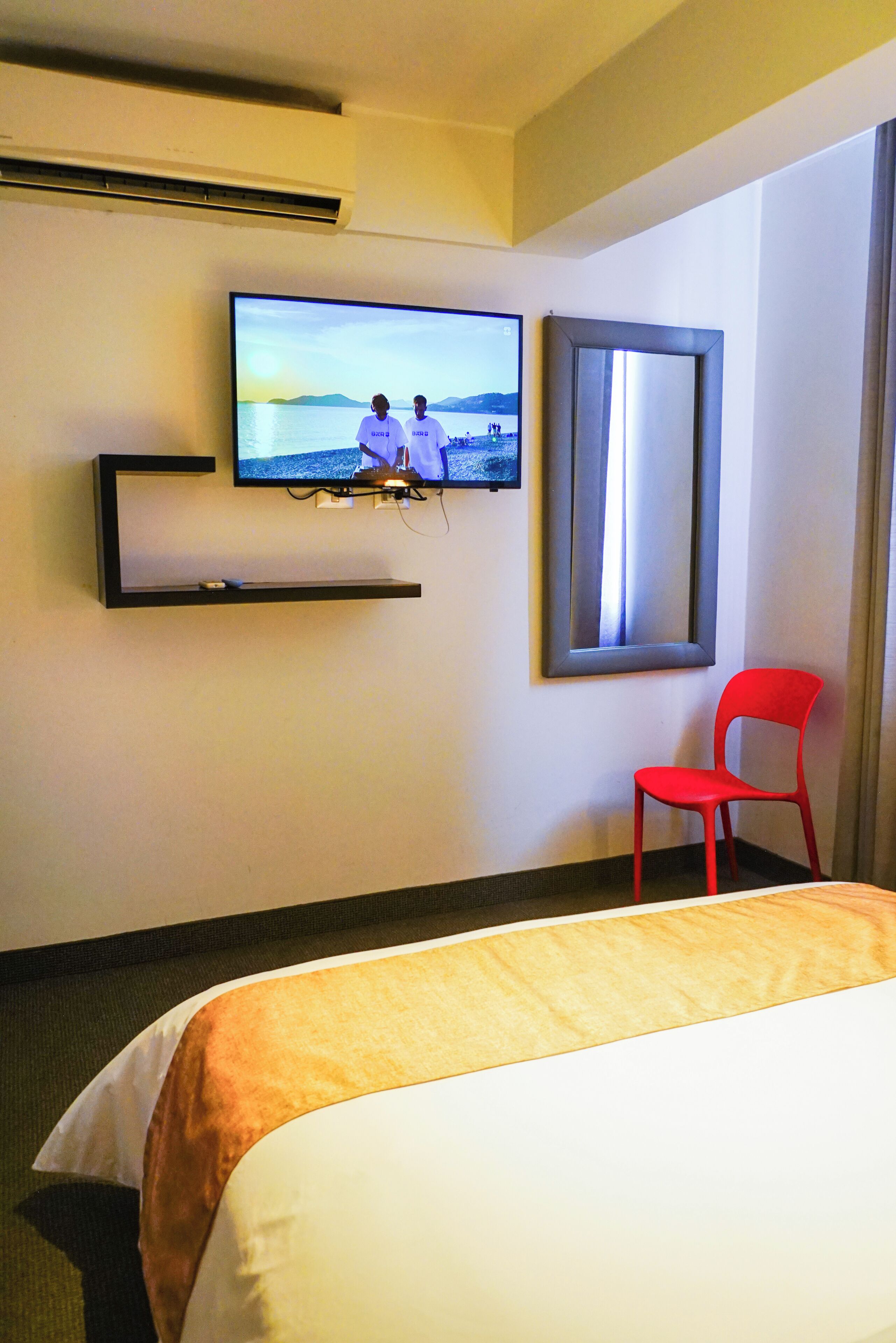 A hotel room with a bed, a television mounted on the wall, a red chair, and a framed mirror.