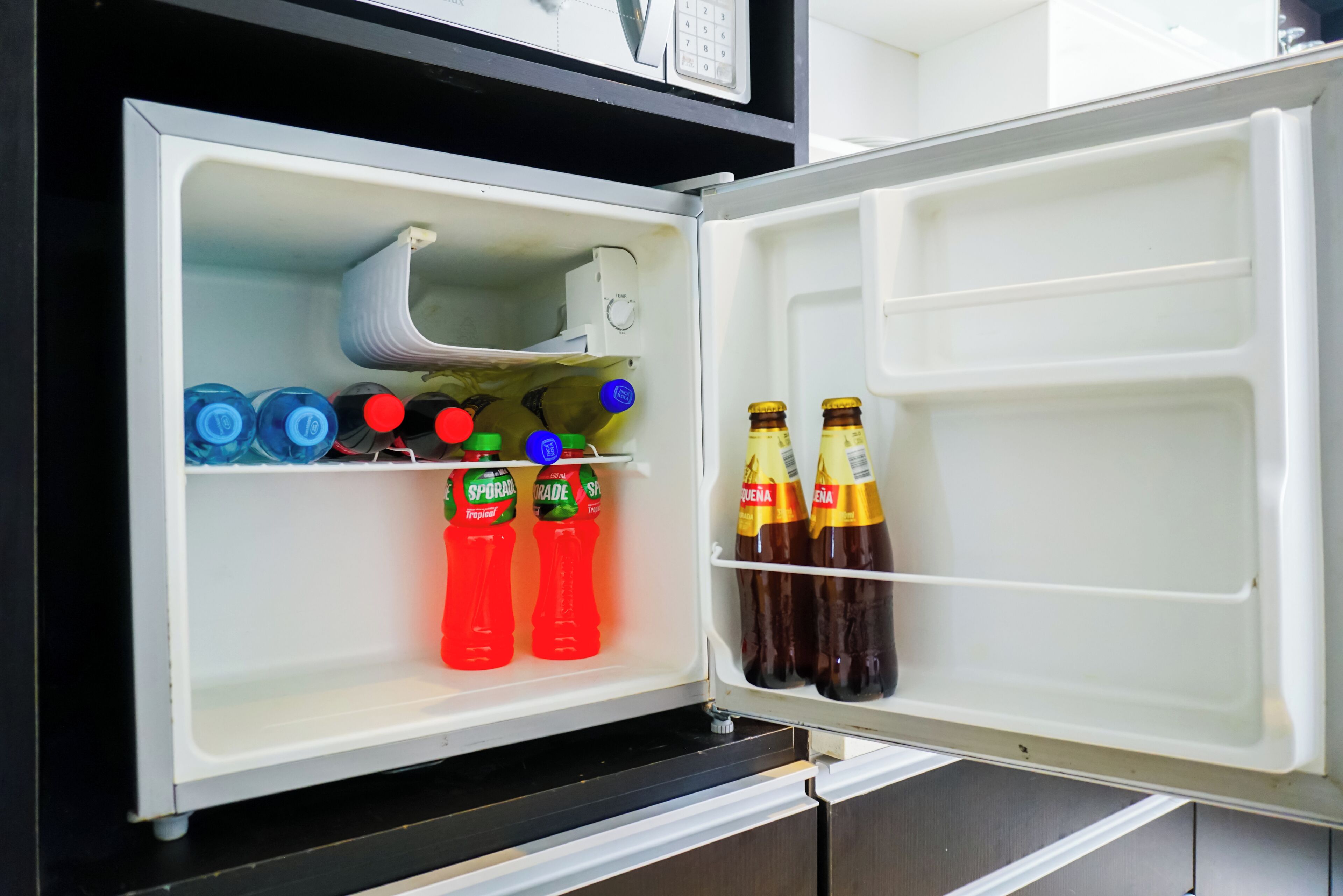 Ein offener Kühlschrank mit Regalen, die mit Getränkeflaschen und einer Mikrowelle gefüllt sind.