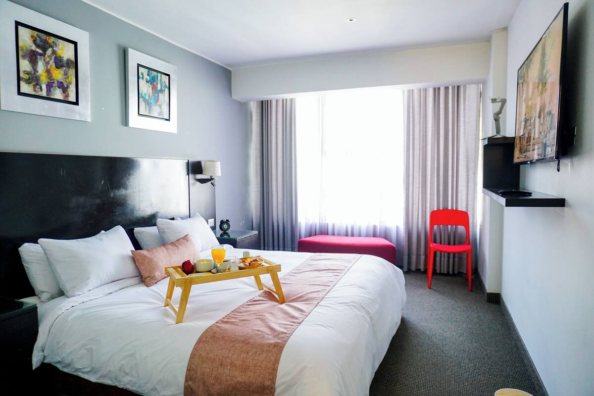 Quarto de hotel com uma cama grande, uma mesinha de cabeceira com abajur, uma cadeira vermelha e uma janela com cortinas.