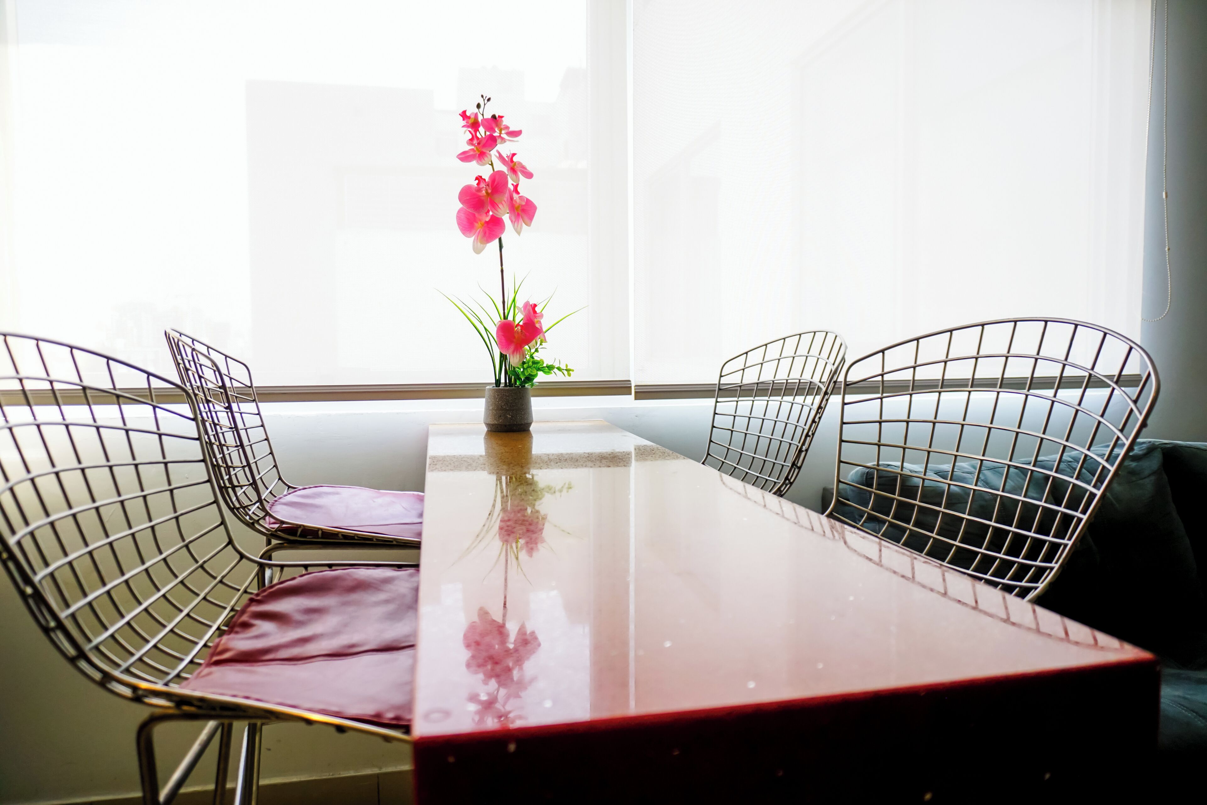 A dining table with wire mesh chairs, a vase with pink flowers, and a view of the outside through a window.