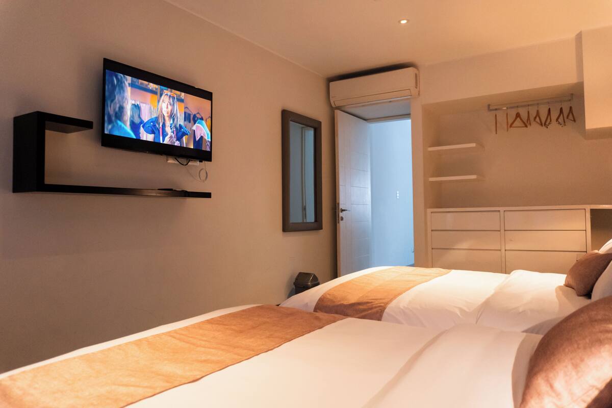 Quarto de hotel com televisão na parede, cama com roupa de cama branca e castanha, guarda-roupa com roupas penduradas e porta de acesso a uma casa de banho.