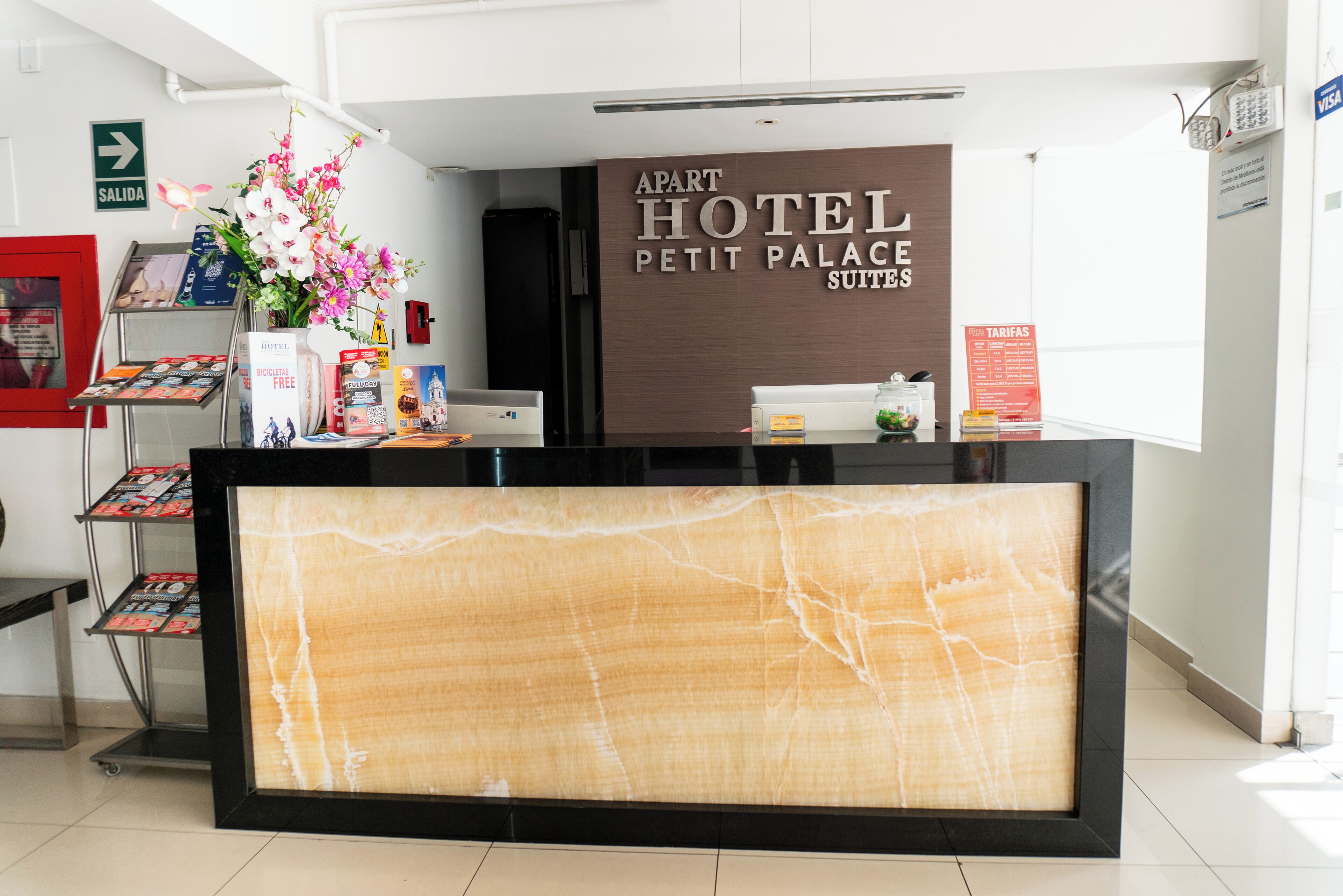 Una recepción con un cartel que dice "Apart Hotel Petit Palace Suites", un arreglo floral y una pantalla digital que muestra un patrón similar al mármol.