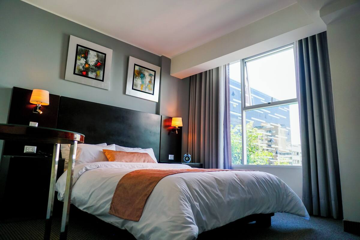 Quarto de hotel com cama, duas luminárias de cabeceira, mesa de cabeceira, janela com cortinas e duas fotografias emolduradas na parede.