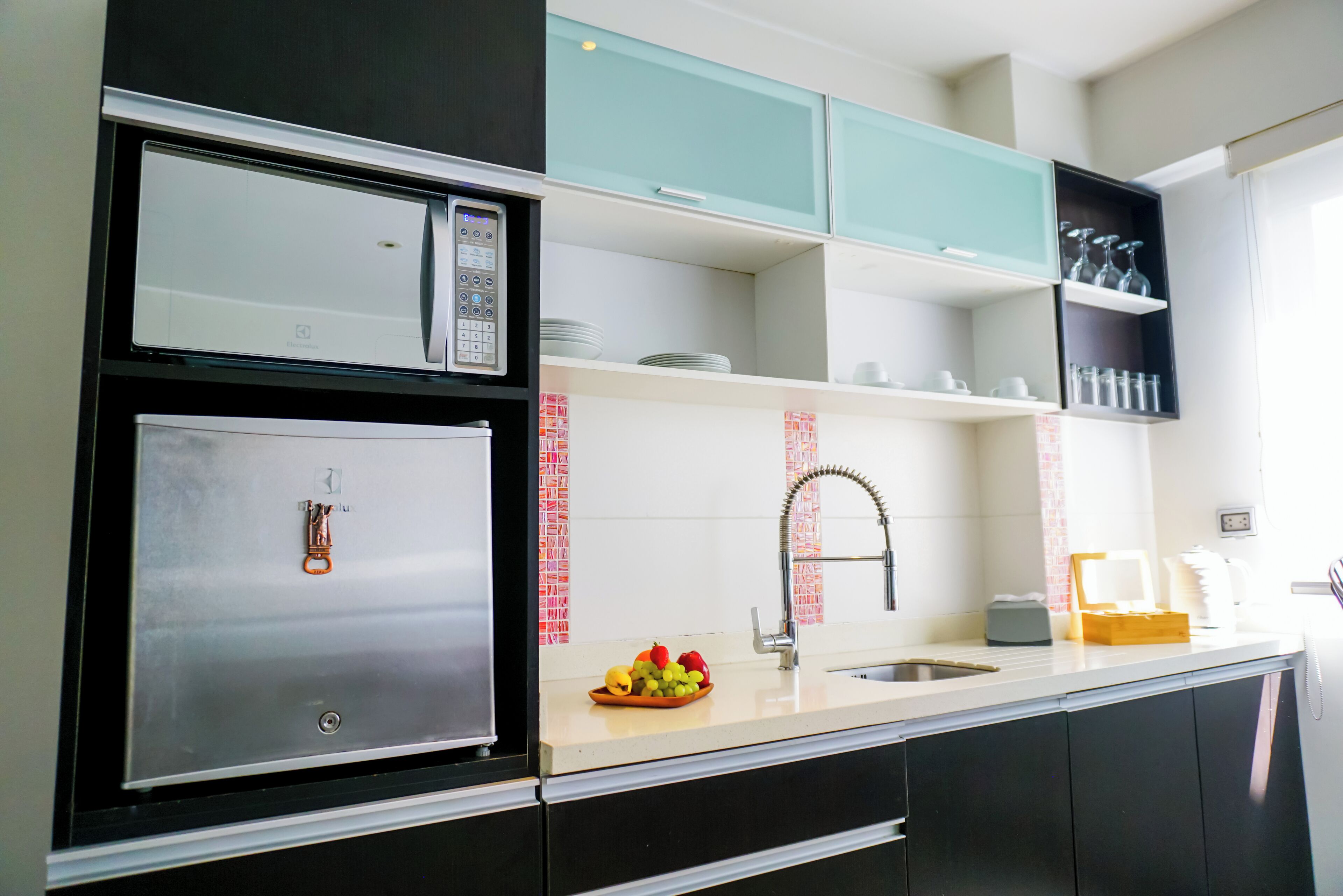 A modern kitchen with a stainless steel refrigerator, a microwave, and a sink with a faucet. There are cabinets above the counter and a fruit bowl on the counter.