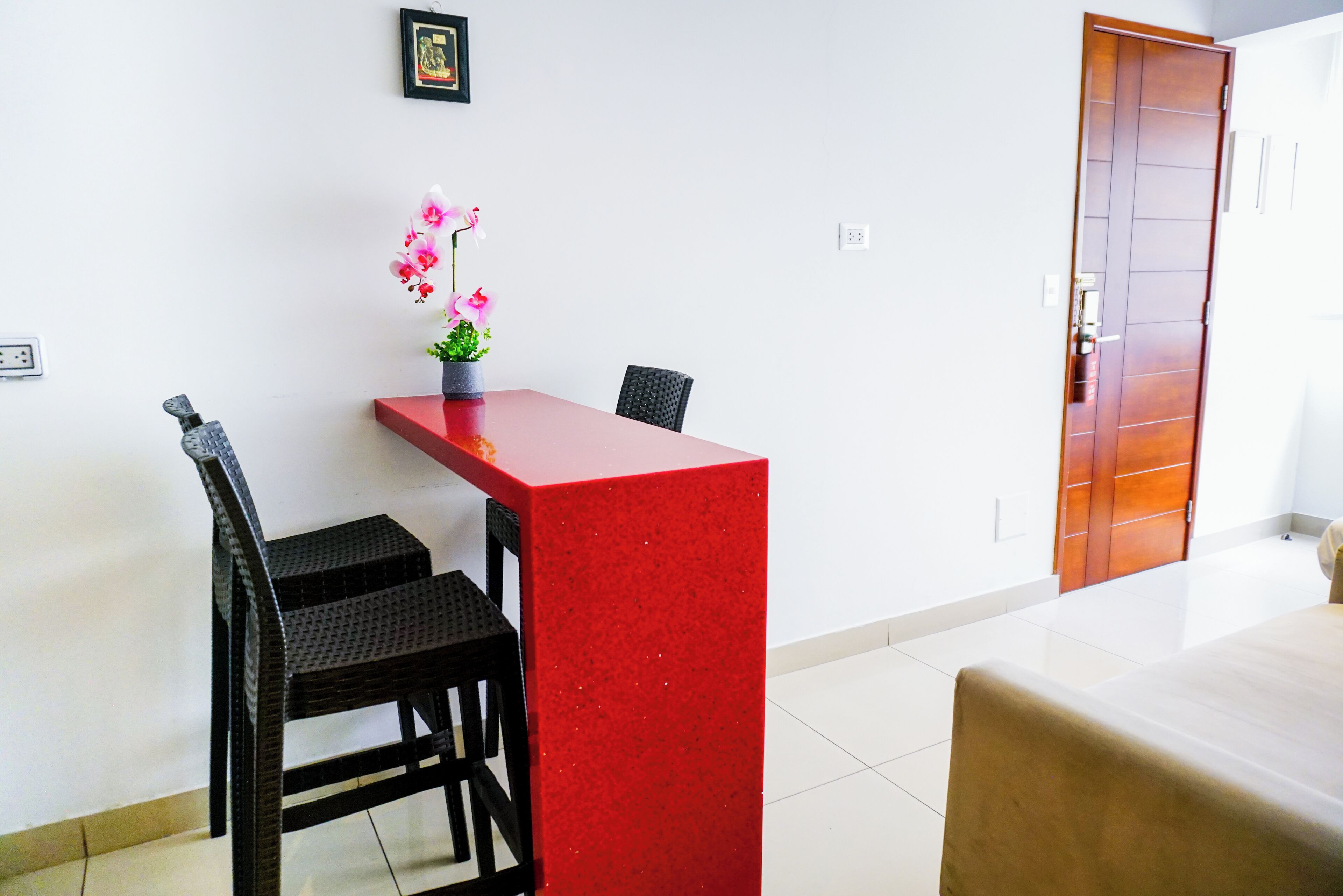 A compact hotel room with a red accent wall, a small dining table with two chairs, a potted orchid, and a wooden door.