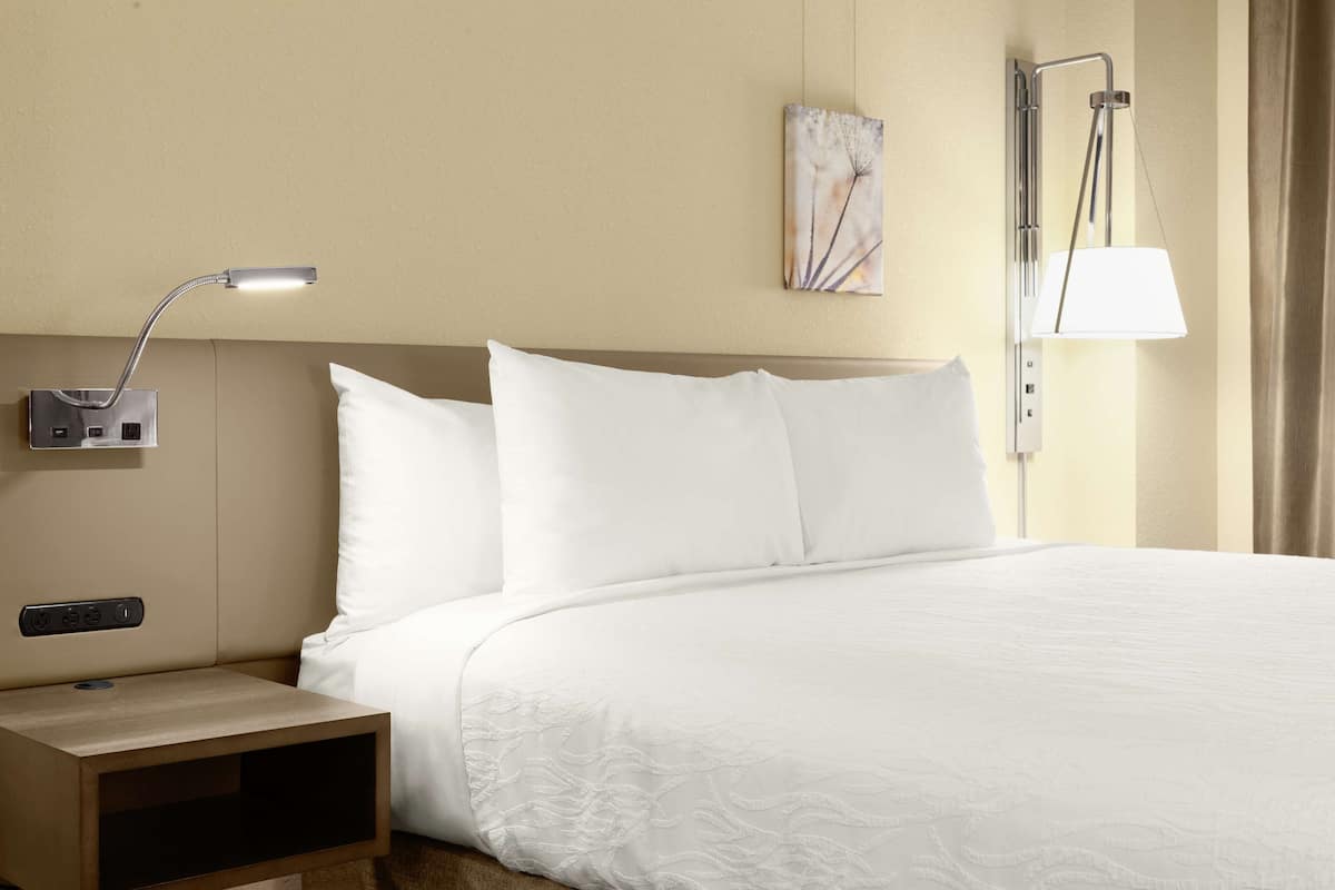A hotel room with a bed, a nightstand, a wall-mounted lamp, and a painting.