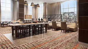 Una moderna hall d'hotel con zona pranzo centrale, posti a sedere e ampie finestre che lasciano entrare la luce naturale.