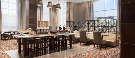 Un moderno vestíbulo de hotel con una zona de comedor central, arreglos de asientos y grandes ventanales que proporcionan luz natural.