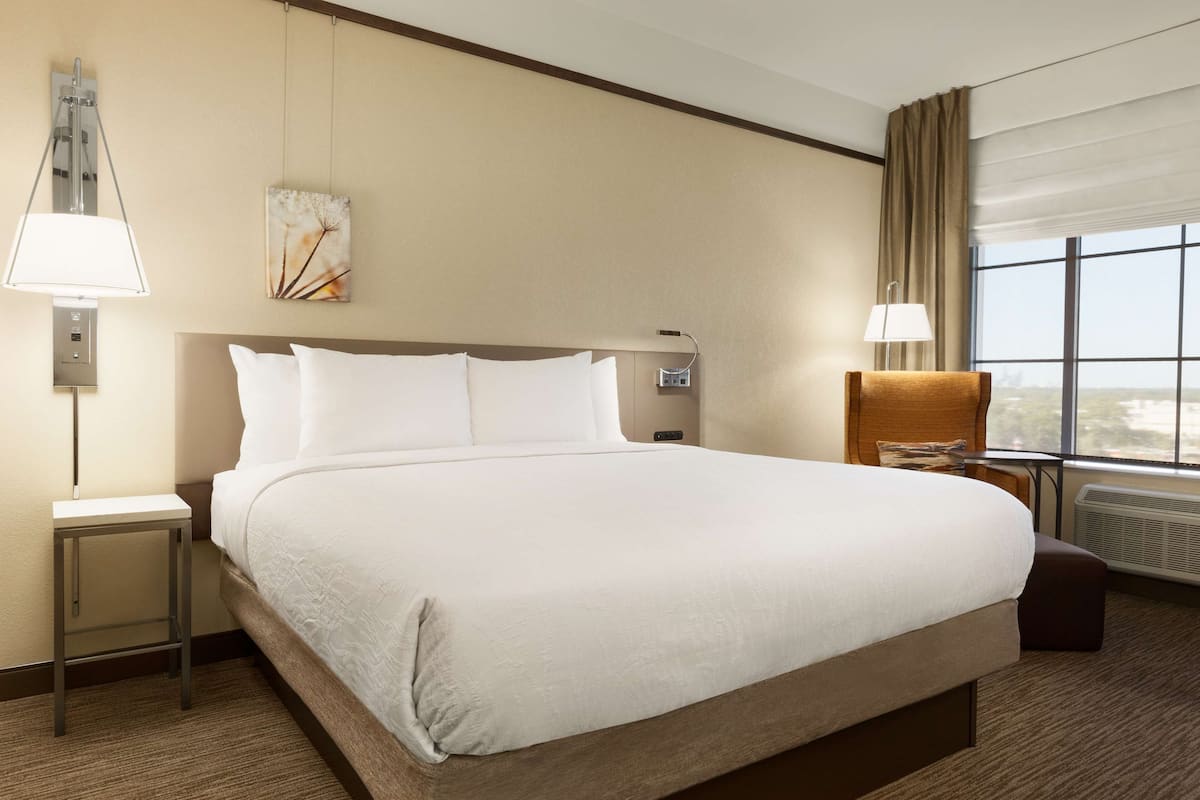 A hotel room with a large bed, two bedside tables, a nightstand, a window with curtains, and a wall-mounted lamp.