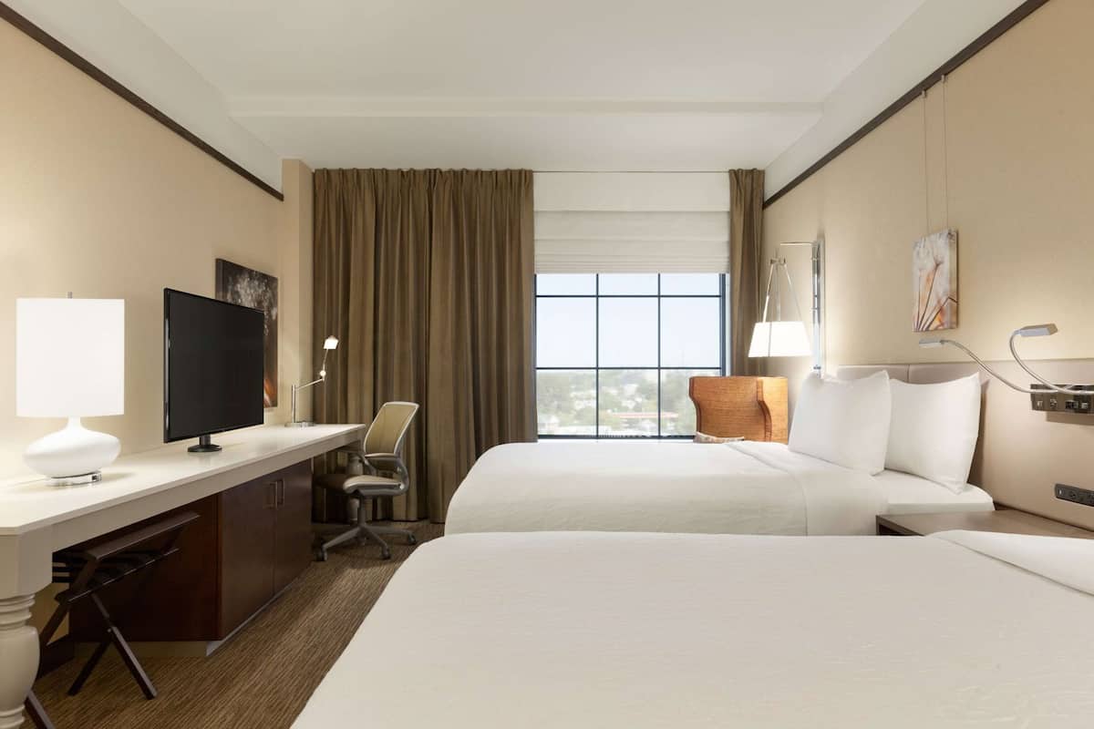 A hotel room with two beds, a desk with a TV, a chair, a lamp, and a window with curtains.