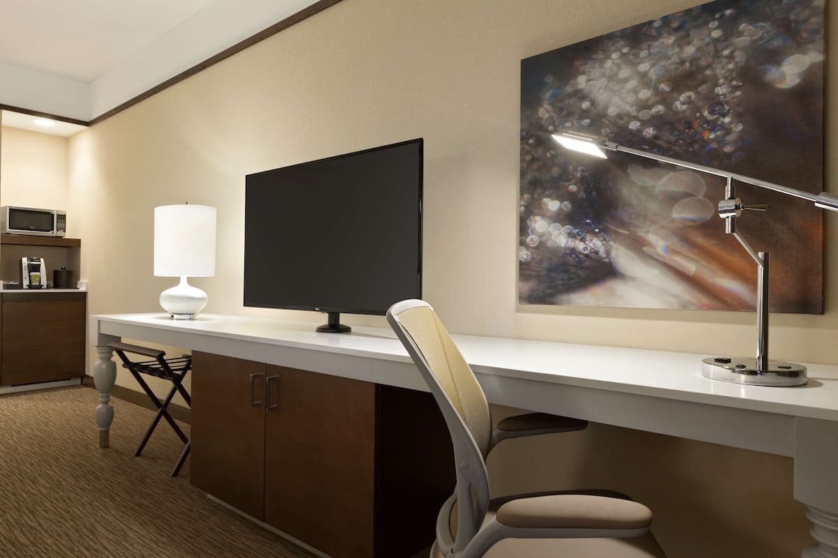 A hotel room with a desk, a flat-screen TV, a lamp, a microwave, and a painting on the wall.