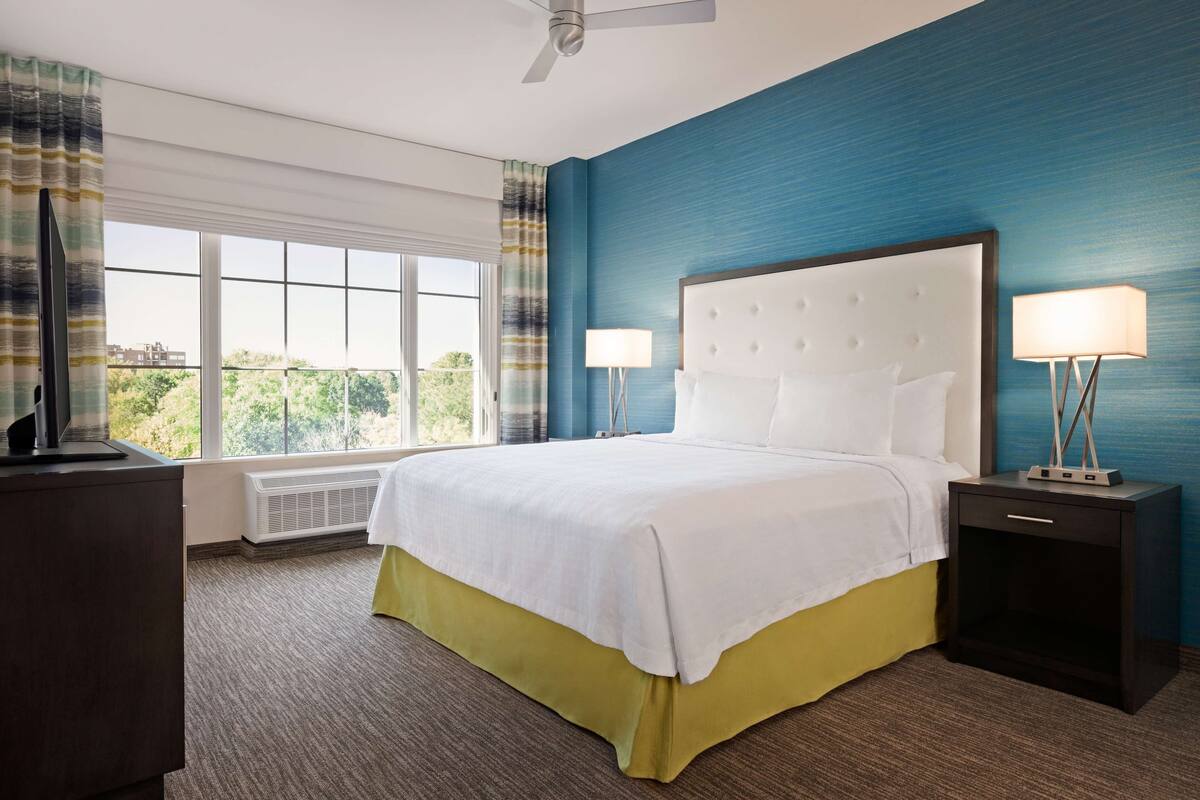 A hotel room with a large bed, two bedside tables with lamps, a ceiling fan, and a view of greenery outside.
