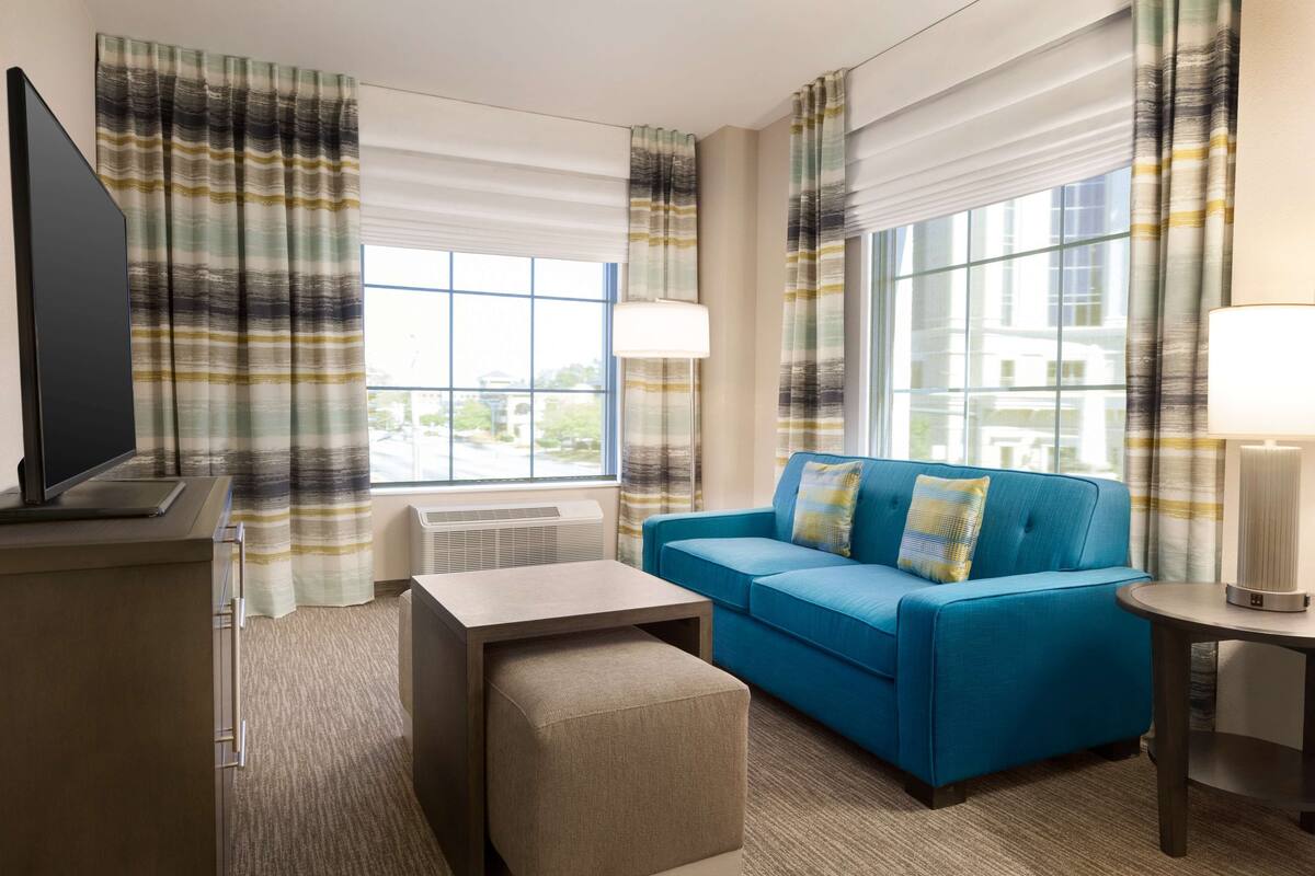 A hotel room with a blue sofa, a TV, a lamp, and large windows with curtains.