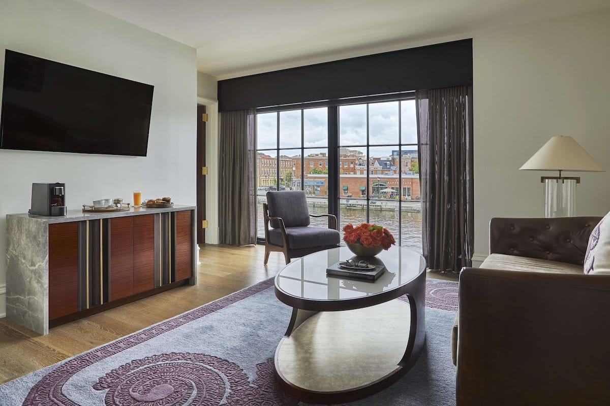A modern living room with a flat-screen TV, a coffee machine, a sofa, a chair, and a large window with a city view.