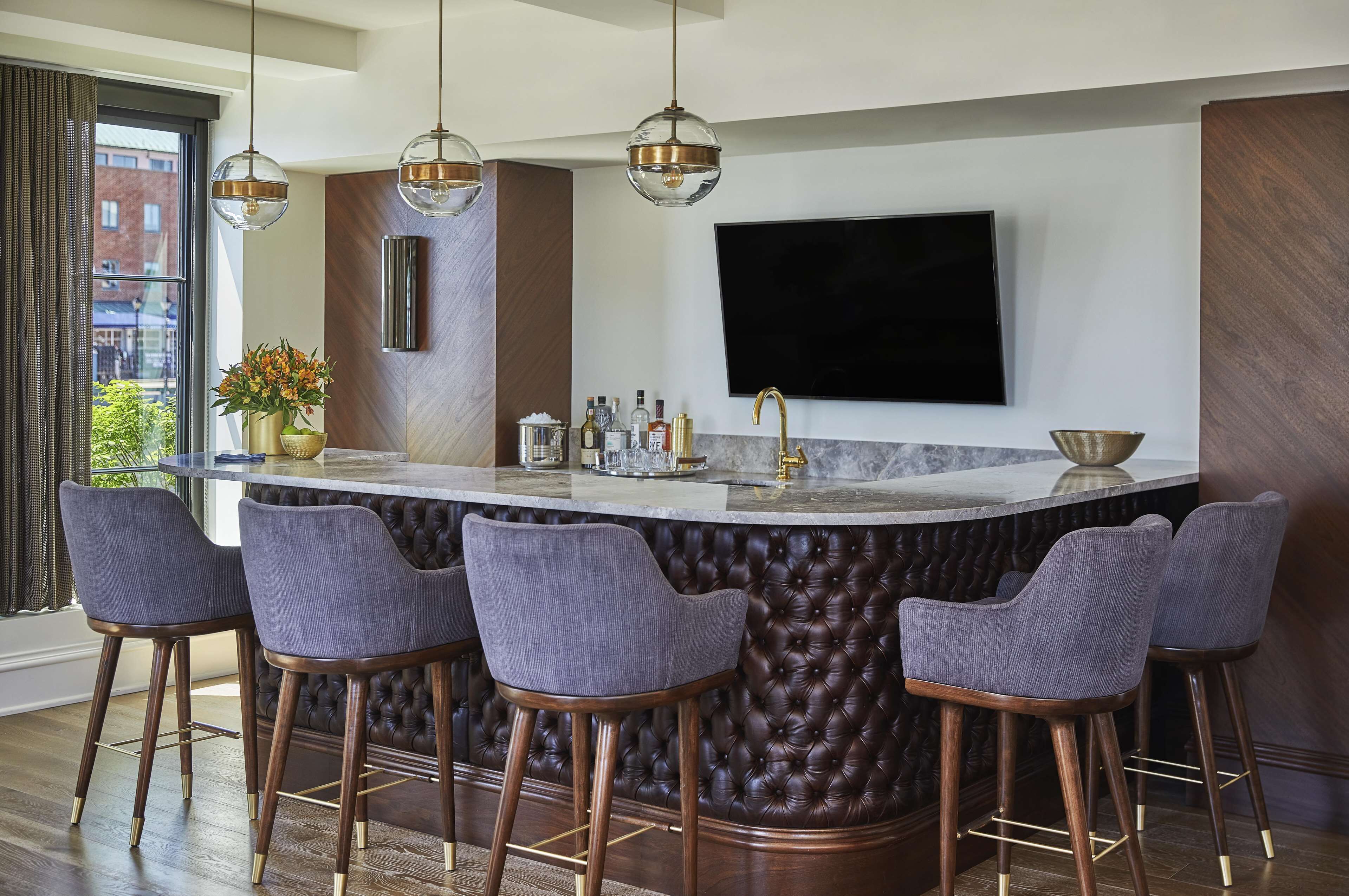 Una cocina moderna con barra, taburetes, un televisor grande y un ventanal con cortinas.