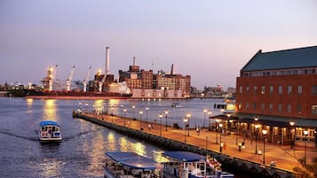 A waterfront promenade with a boat docked on the left, a red brick building on the right, and a large industrial complex in the background.