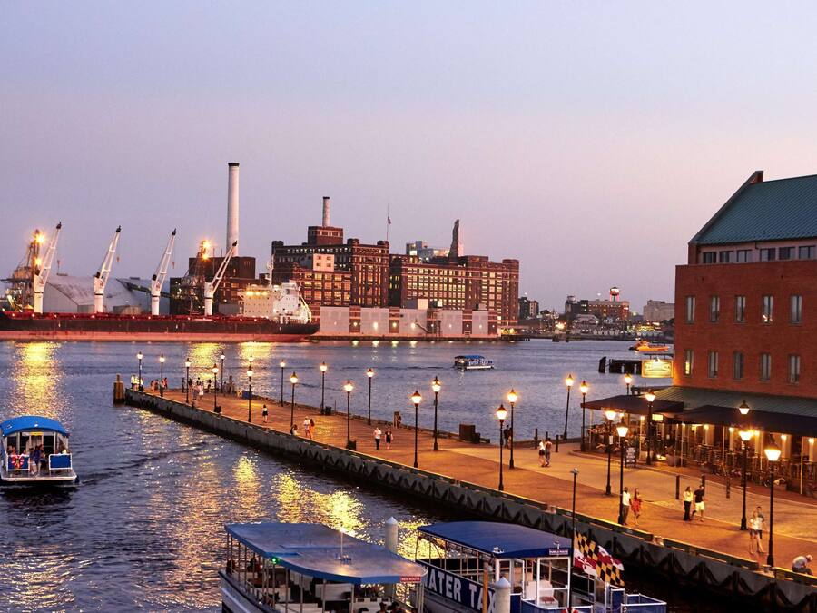 A waterfront promenade with a boat docked on the left, a red brick building on the right, and a large industrial complex in the background.