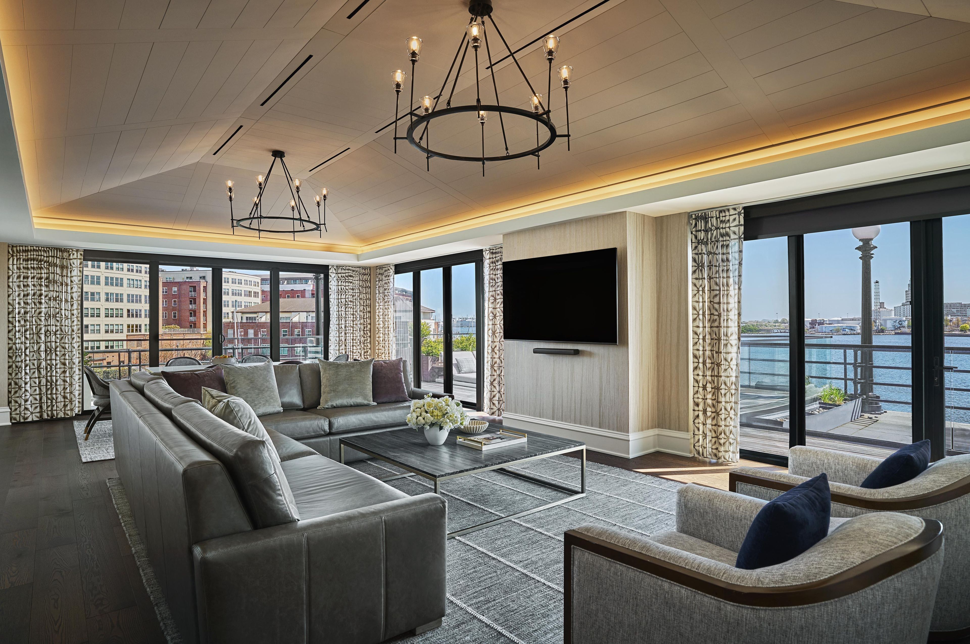 A modern living room with a sectional sofa, a coffee table, and a TV. There are large windows with a view of a cityscape and a body of water.
