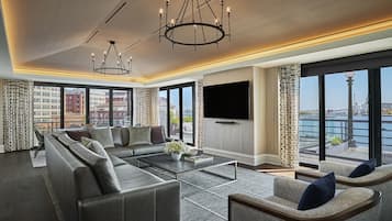 A modern living room with a sectional sofa, a coffee table, and a TV. There are large windows with a view of a cityscape and a body of water.