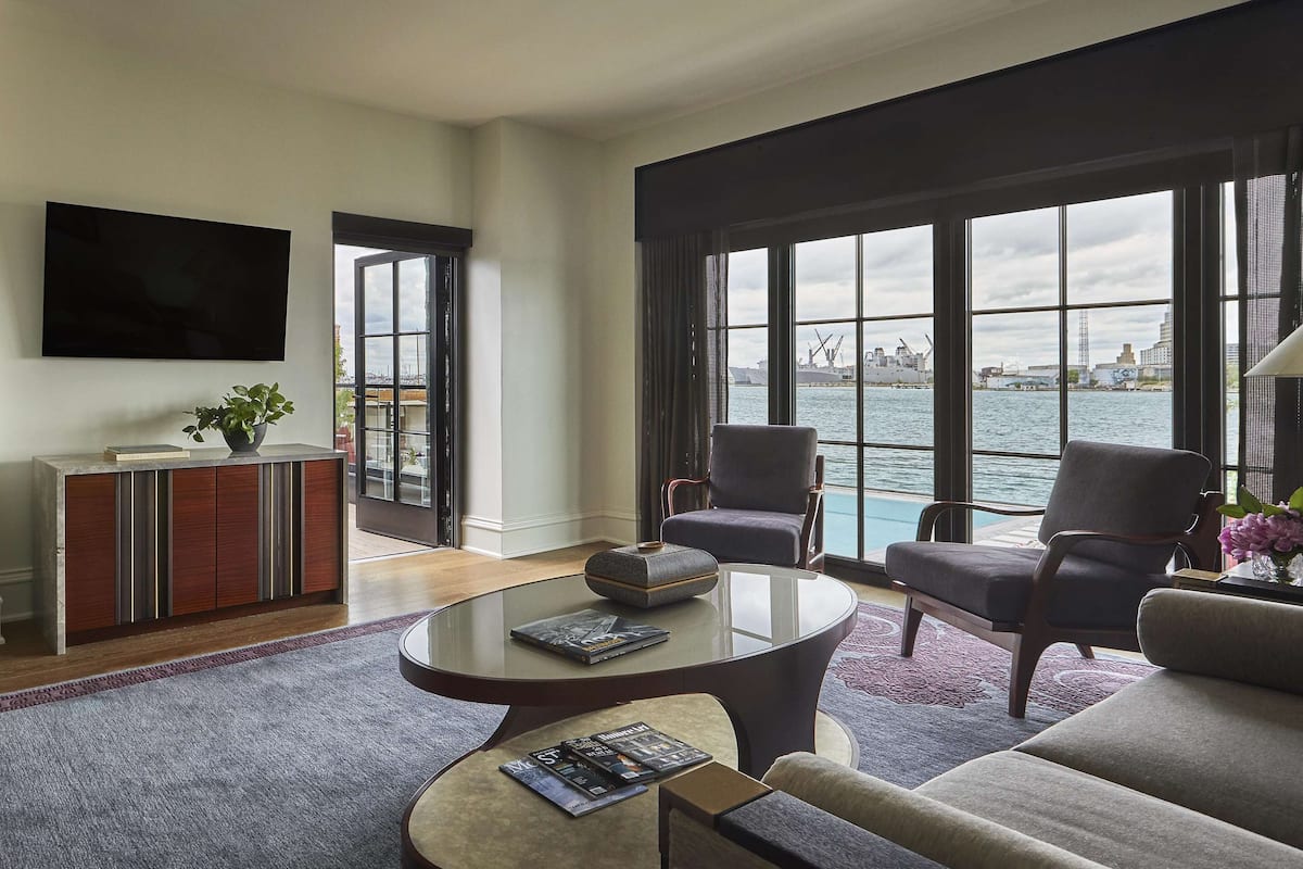 A hotel room with a large window offering a view of the water and cityscape, a flat-screen TV mounted on the wall, a modern sofa, a coffee table, and a side table with a magazine.