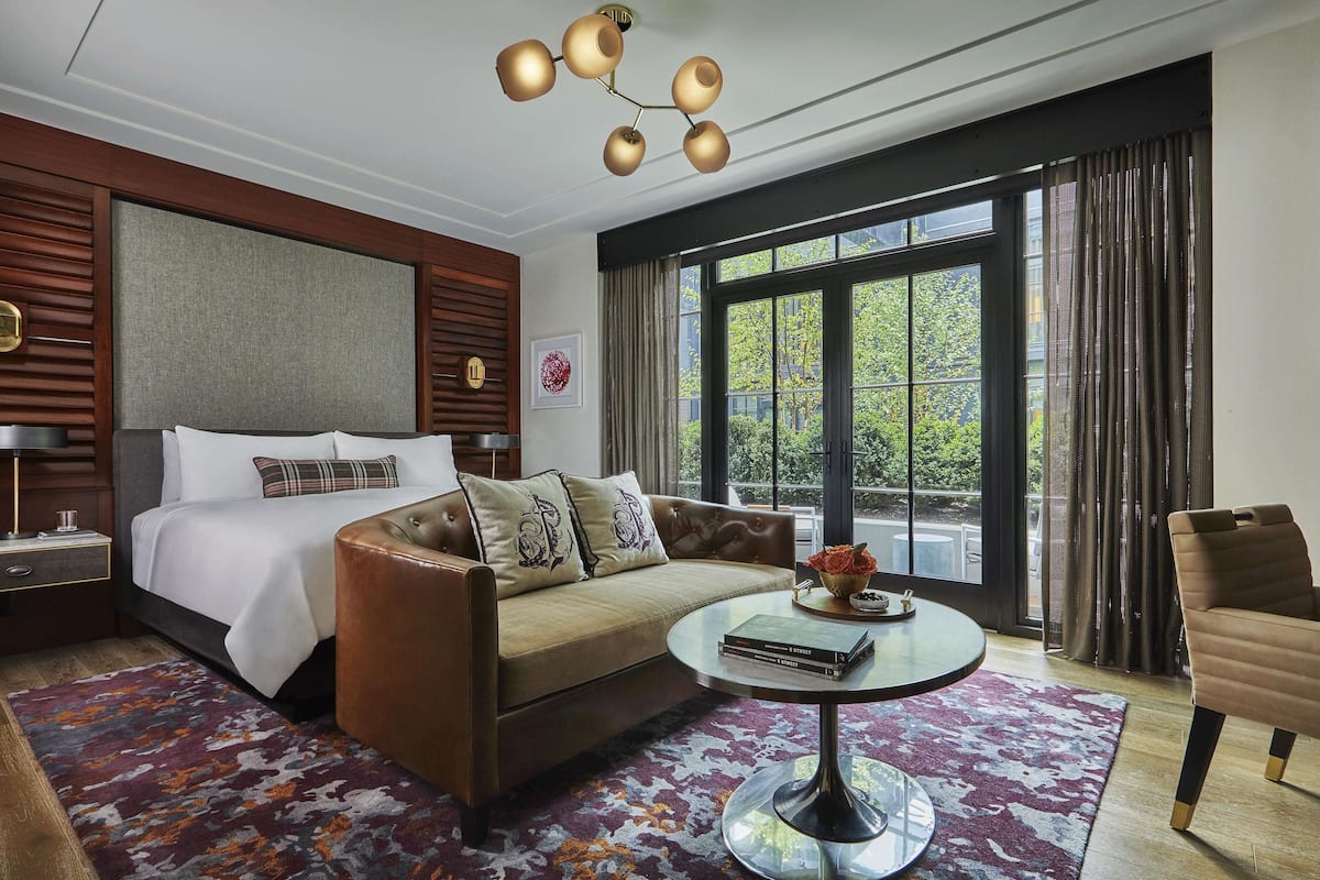 A modern hotel room with a large bed, a sofa, a coffee table, and a view of greenery outside.