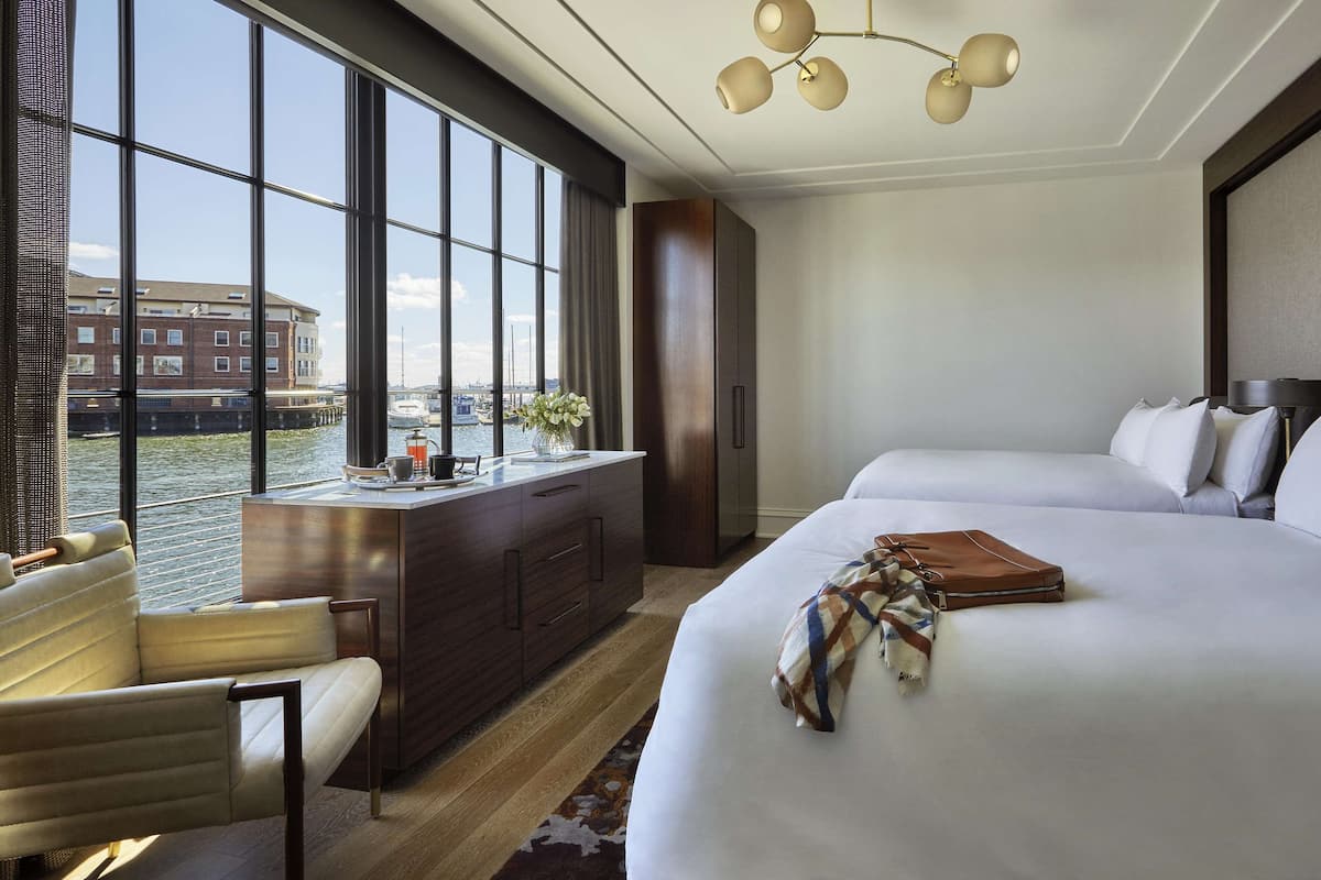 A hotel room with two beds, a desk with a chair, a view of a canal, and a large window.