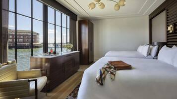 A hotel room with two beds, a desk with a chair, a view of a canal, and a large window.