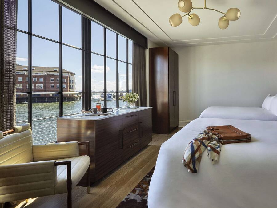 A hotel room with two beds, a desk with a chair, a view of a canal, and a large window.