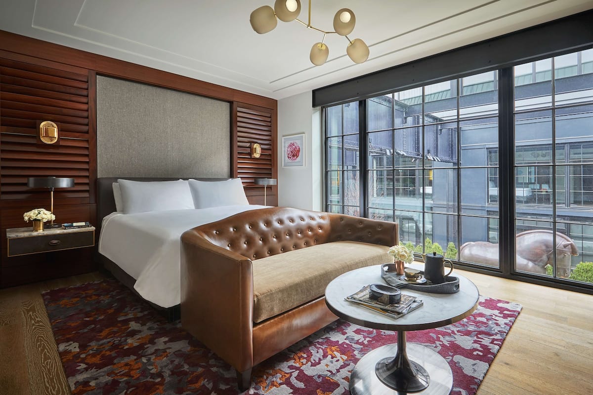 A modern hotel room with a large bed, a sofa, a round coffee table, and a large window offering a view of the city.
