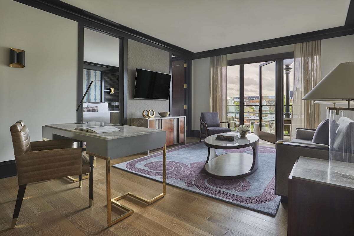 A modern hotel room with a large TV, a dining area, a sofa, and a view of the city.