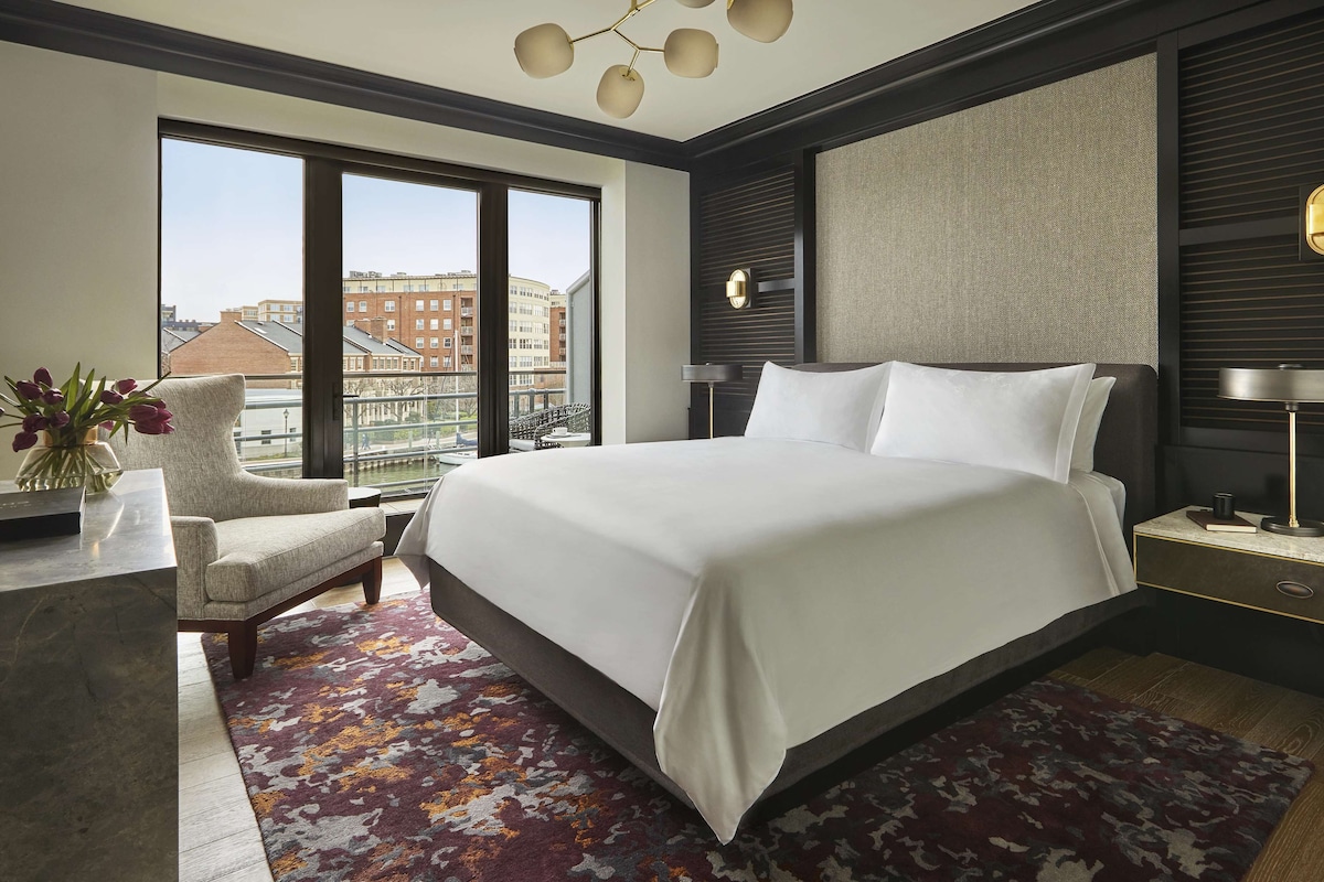 Une chambre d’hôtel moderne dotée d’un grand lit, d’un coin salon avec un fauteuil, d’une table de chevet et offrant une vue sur le paysage urbain grâce à de grandes fenêtres.