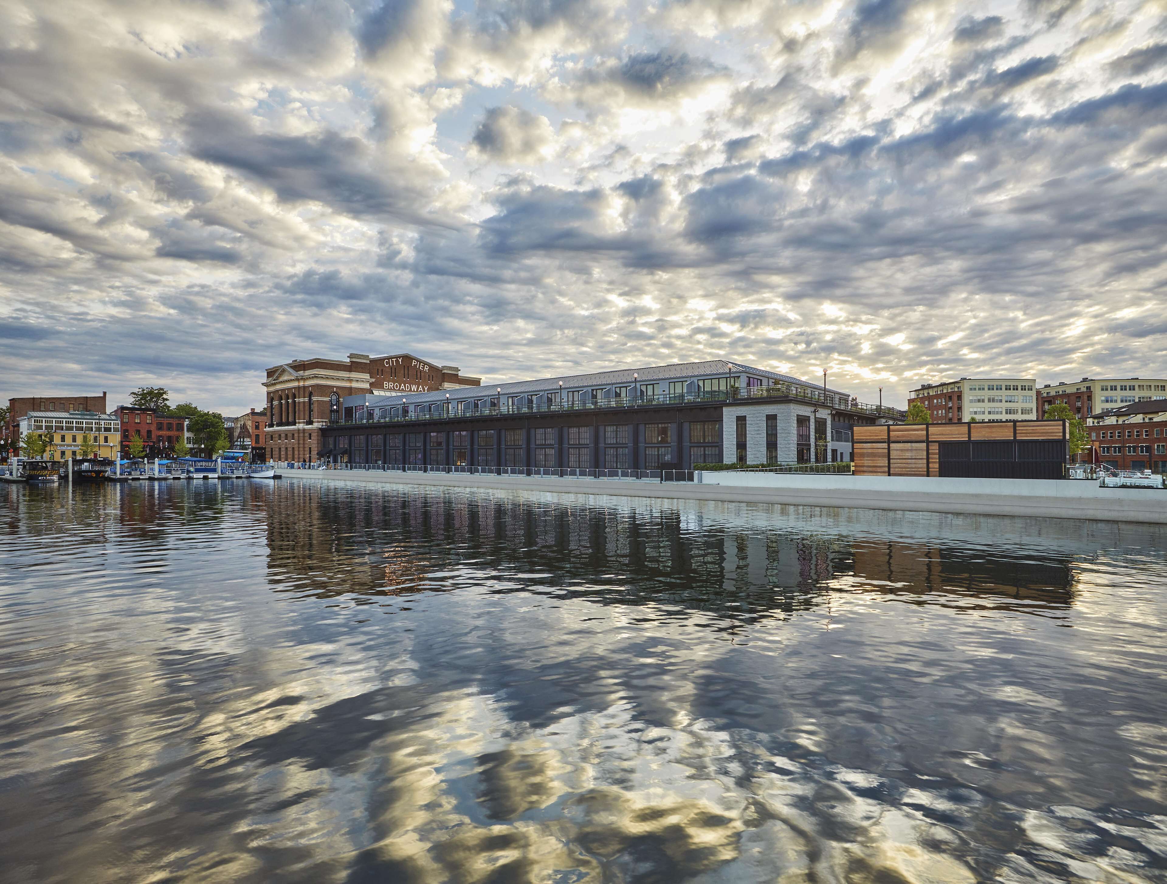 A waterfront building with a glass facade, a reflection in the water, and a city pier in the background.