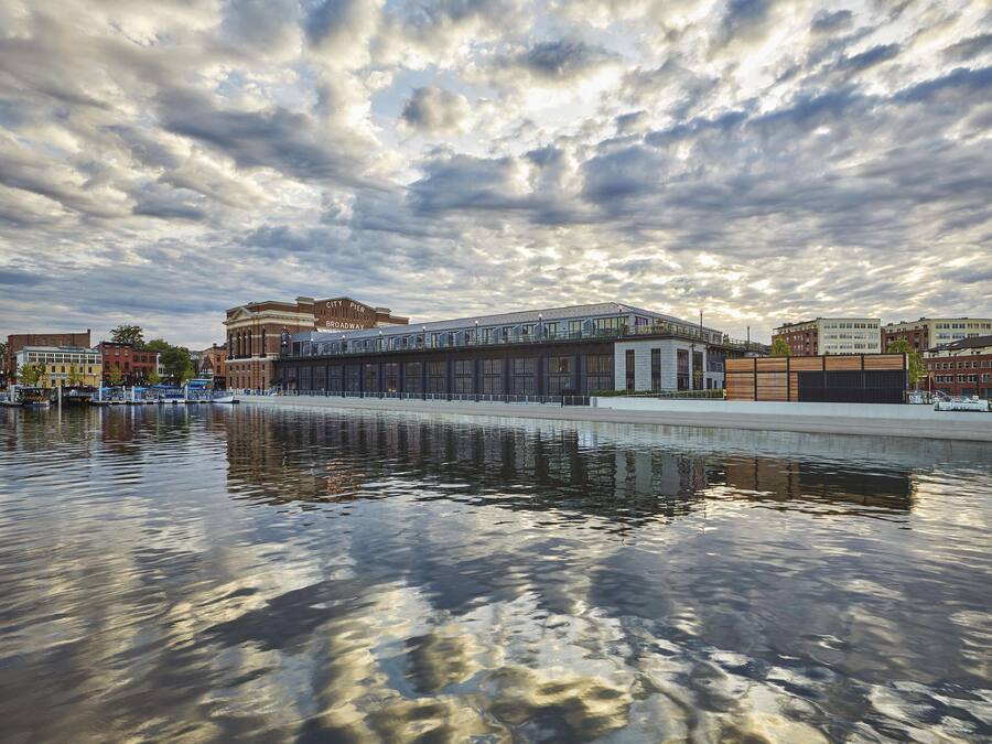 A waterfront building with a glass facade, a reflection in the water, and a city pier in the background.