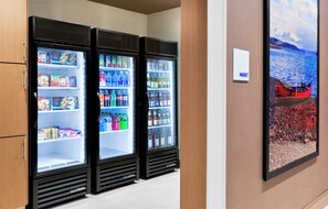 A row of beverage refrigerators stocked with various drinks and snacks, a wall-mounted sign indicating a market, and a framed picture of a boat on water. - Holiday Inn Express & Suites Prosser - Yakima Valley Wine by IHG (Prosser)