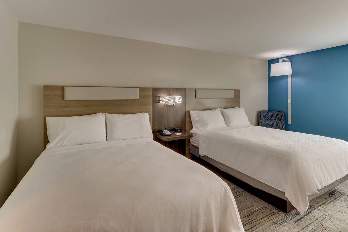 A hotel room with two beds, a nightstand with a phone, and a wall-mounted lamp.