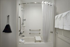 Een badkamer met een badkuip, douche, grepen, een handdoekhouder en een zwarte tas die aan een haak hangt.