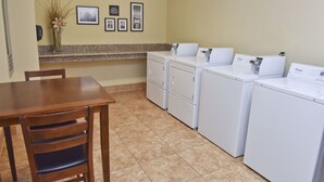 A laundry area with a wooden table and chairs, a vase with dried plants, and a row of white washing machines.