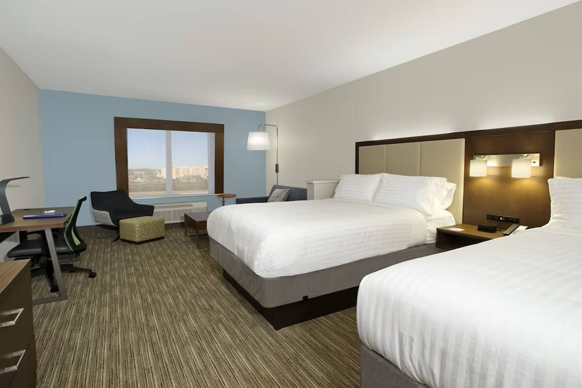 A hotel room with two beds, a desk, a chair, a small table, and a window with a view.
