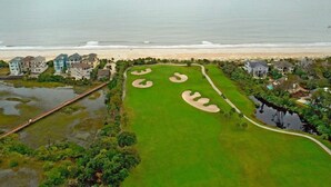 Vista aérea de un campo de golf costero con búnkeres, un puente y propiedades frente a la playa.