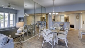 Una sala de estar moderna con un sofá gris, una mesa de comedor con sillas blancas y una cocina visible a través de una pared de vidrio.